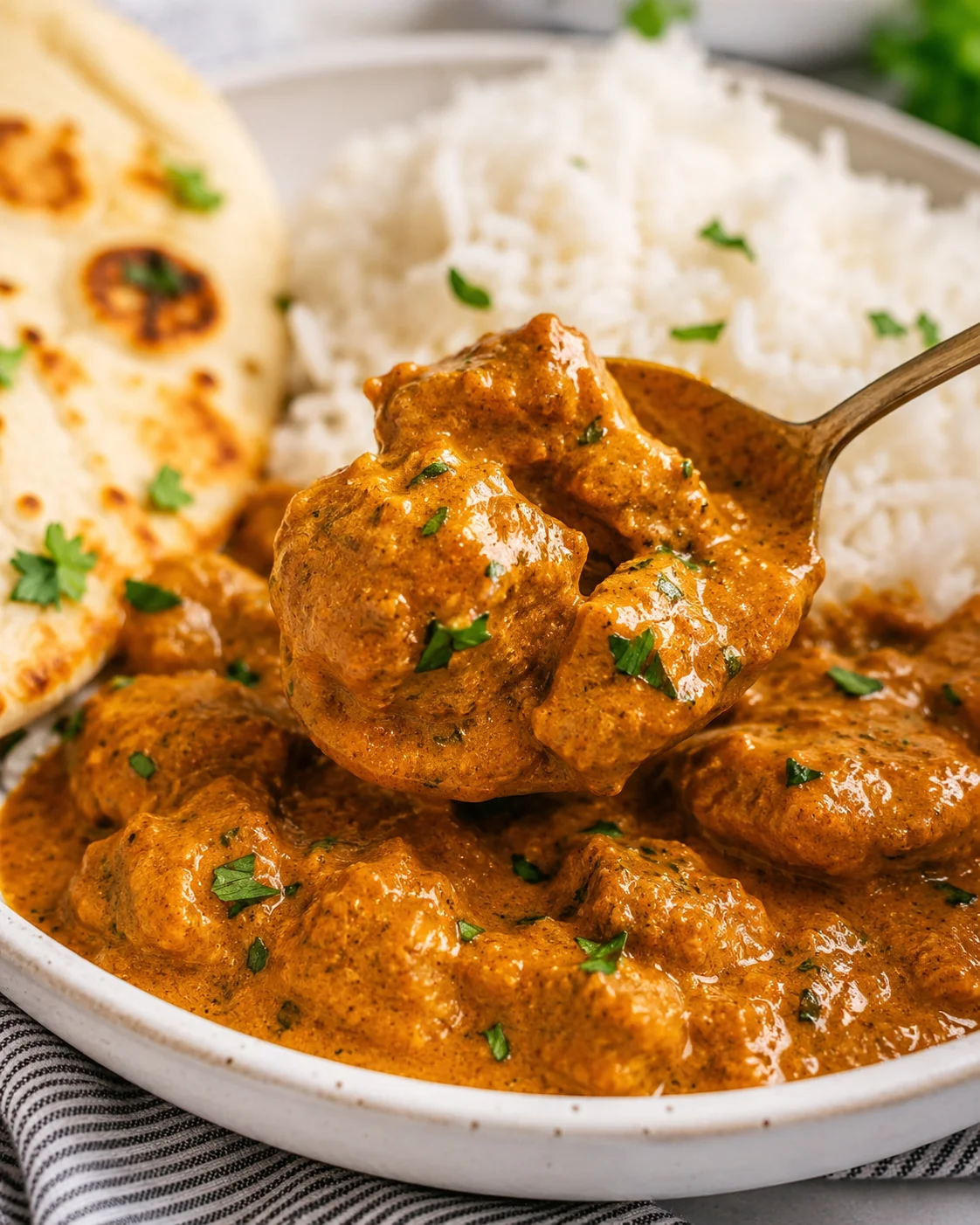 Spoonful of butter chicken served with white rice and naan in the background