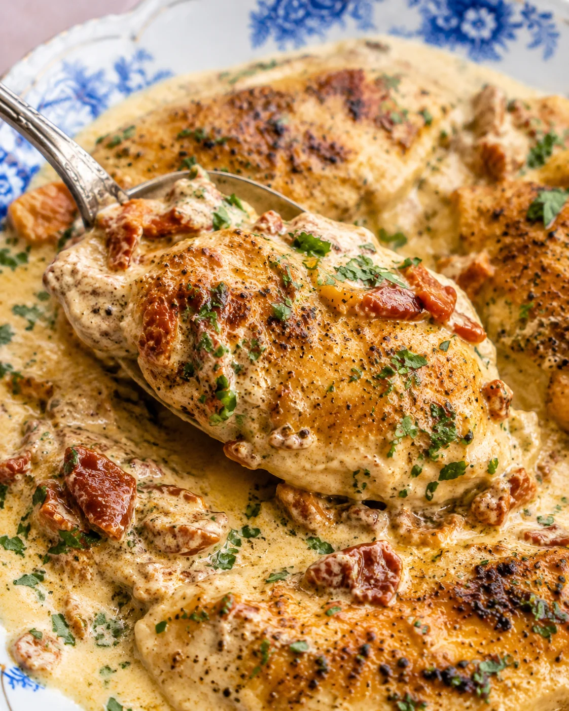 Spoon lifting a seared chicken breast from creamy sun-dried tomato sauce