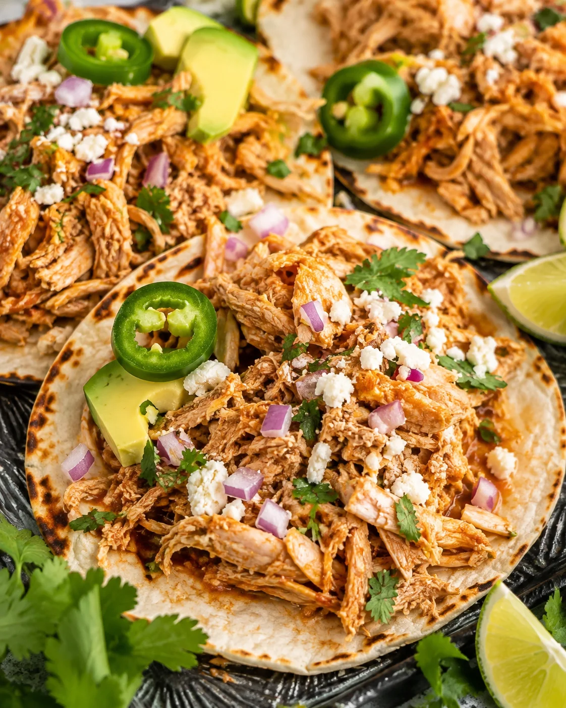 Healthy crockpot chicken tacos on tortillas topped with avocado, jalapeño, red onion and crumbled cheese