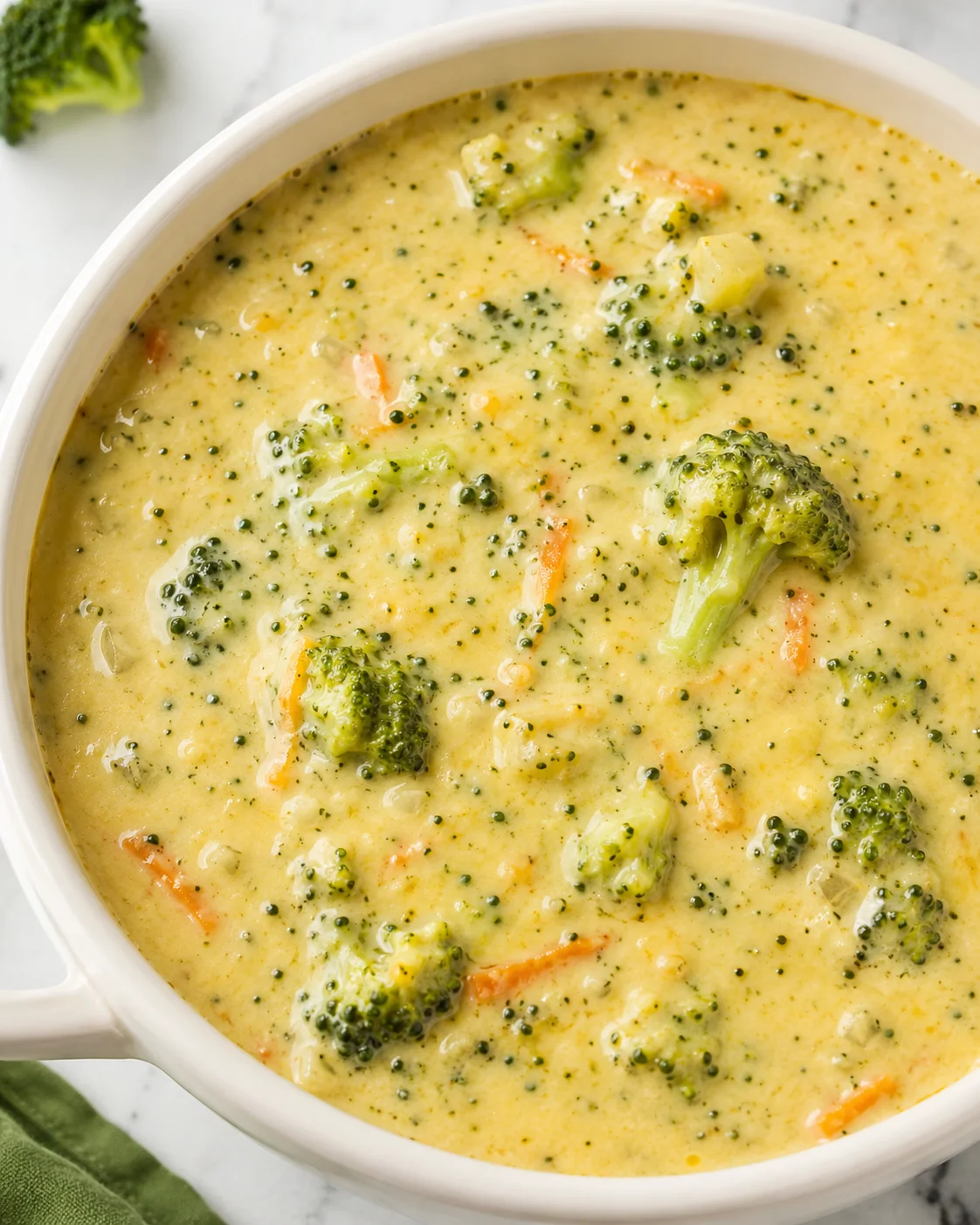 Overhead close-up of High Protein Broccoli Cheddar Soup showing creamy broth and broccoli florets in a white bowl.