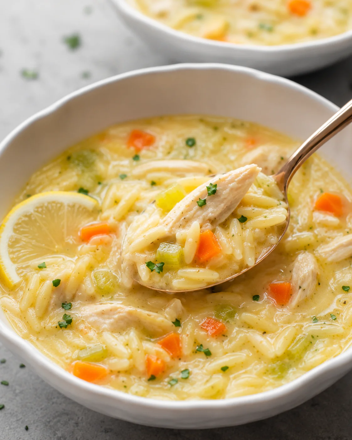 Spoon lifting a bite of lemon chicken orzo soup with chicken, orzo and a lemon slice visible in the bowl